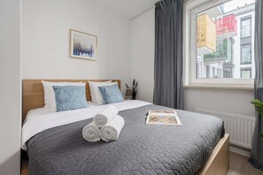 Une chambre d’hôtel avec un lit, des oreillers, une serviette roulée, un magazine, une fenêtre avec des rideaux et une vue sur la ville.