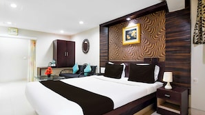 A hotel room with a large bed, a desk, a chair, a small table with a vase of flowers, a clock on the wall, and a patterned wall panel. - Collection O Railway Station (Raipur)