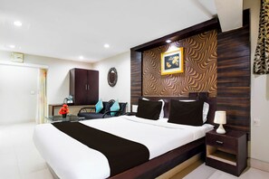 A hotel room with a large bed, a desk, a chair, a small table with a vase of flowers, a clock on the wall, and a patterned wall panel. - Collection O Railway Station (Raipur)