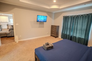 A bedroom with a bed, a television on the wall, a dresser, and a view of the kitchen. - AAccommodation's Corinth Home: Med Center South (Houston)