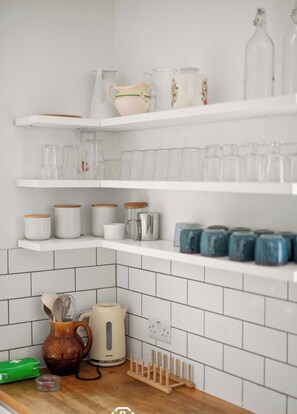 White L-shaped open shelves holding neatly organized glasses, mugs, and jars sit above a wooden countertop with a kettle and a white subway tile backsplash. - Mildred House, Watford (Hertfordshire)