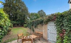 A tiled patio area features a small set of wooden outdoor furniture arranged next to an ivy-covered brick wall with a light blue wooden gate that opens into a grassy backyard. - Mildred House, Watford (Hertfordshire)