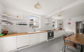 A bright, minimalist kitchen features long white cabinetry with wooden countertops, white subway tile backsplashes, and a large wooden dining table with matching chairs. - Mildred House, Watford (Hertfordshire)