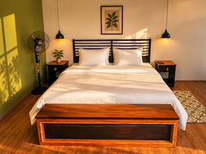 A neatly made bed with a wooden headboard, two bedside tables with lamps, a potted plant, and a framed botanical print on the wall.