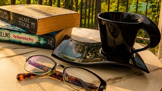 Un tavolo con libri, una tazza di caffè e bicchieri su un balcone affacciato su un paesaggio boschivo.