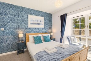 Ein Schlafzimmer mit einem großen Bett, einem Nachttisch, einem Sessel, einem Fenster mit Vorhängen und einer Wand mit Muster.