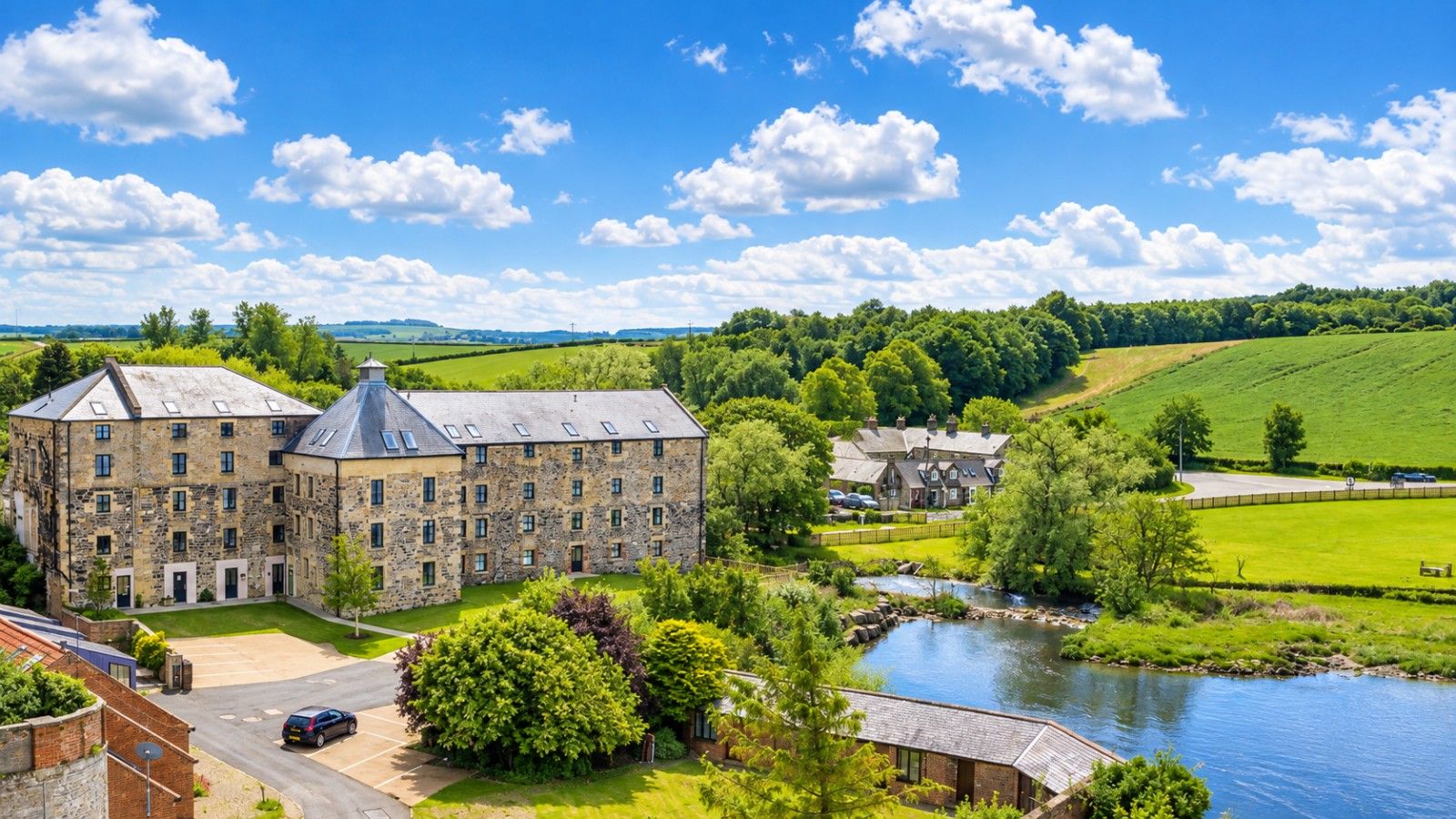 A large stone building with multiple windows and a courtyard, surrounded by lush greenery and a river.