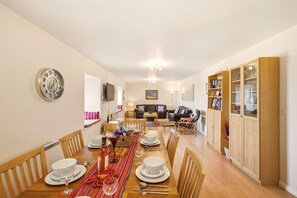 A dining area with a long wooden table set for a meal, flanked by chairs, with a television and seating area in the background. - Bay View @ Waren Mill - North East Escapes (Waren Mill)