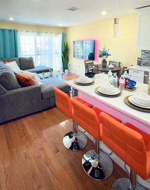 A modern kitchen with a dining area, featuring a bar stool setup, a coffee machine, and a large window with curtains.