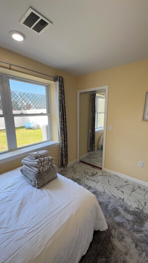 A bedroom with a bed, a window, a mirror, and a view of the outside.