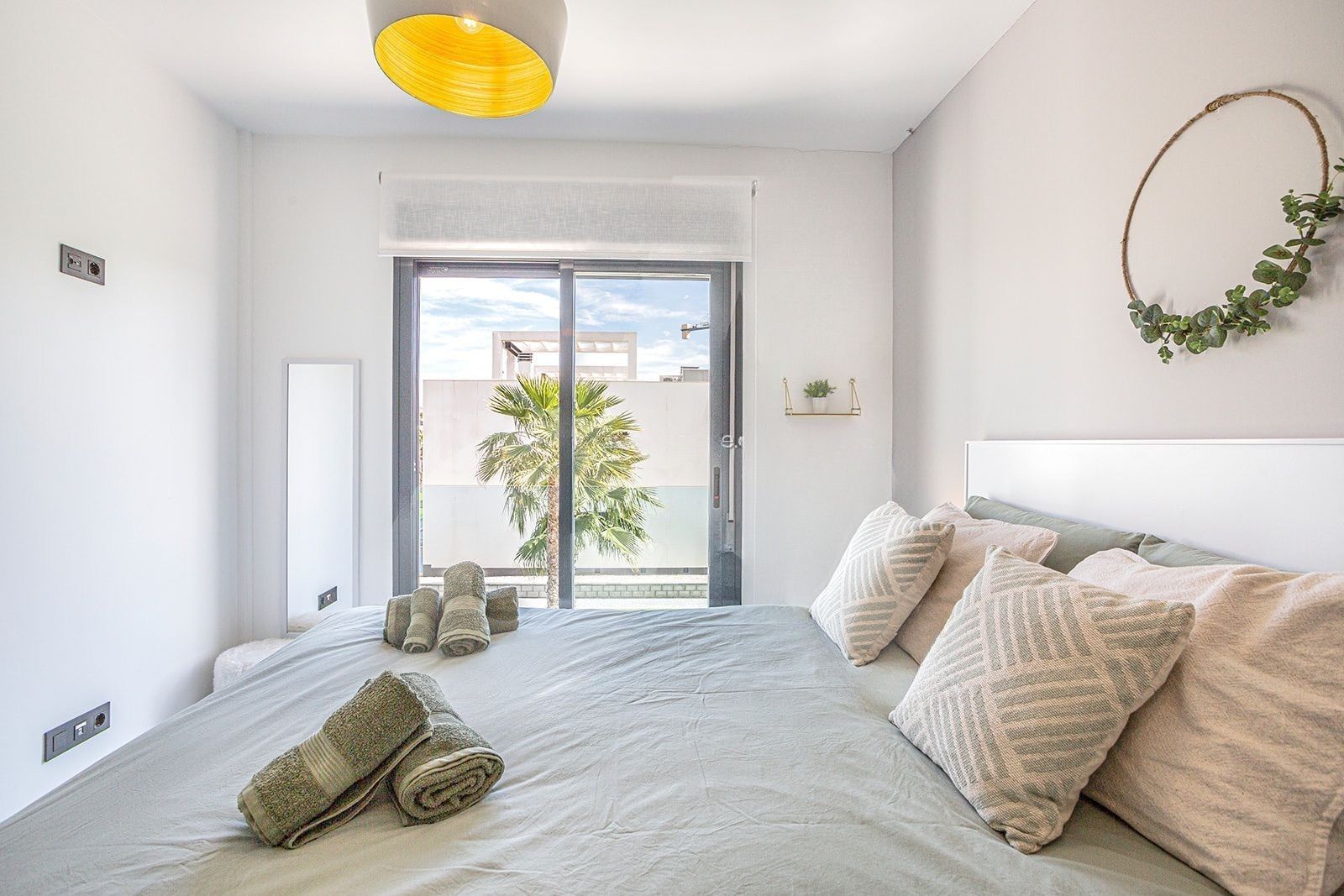 Une chambre d’hôtel avec un lit, une fenêtre donnant sur des palmiers, un luminaire au plafond et une plante fixée au mur.