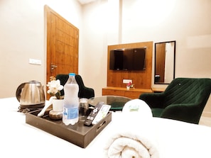 A hotel room with a bed, a wooden wardrobe, a television mounted on the wall, a green armchair, a tray with a kettle, a bottle of water, and a remote control.