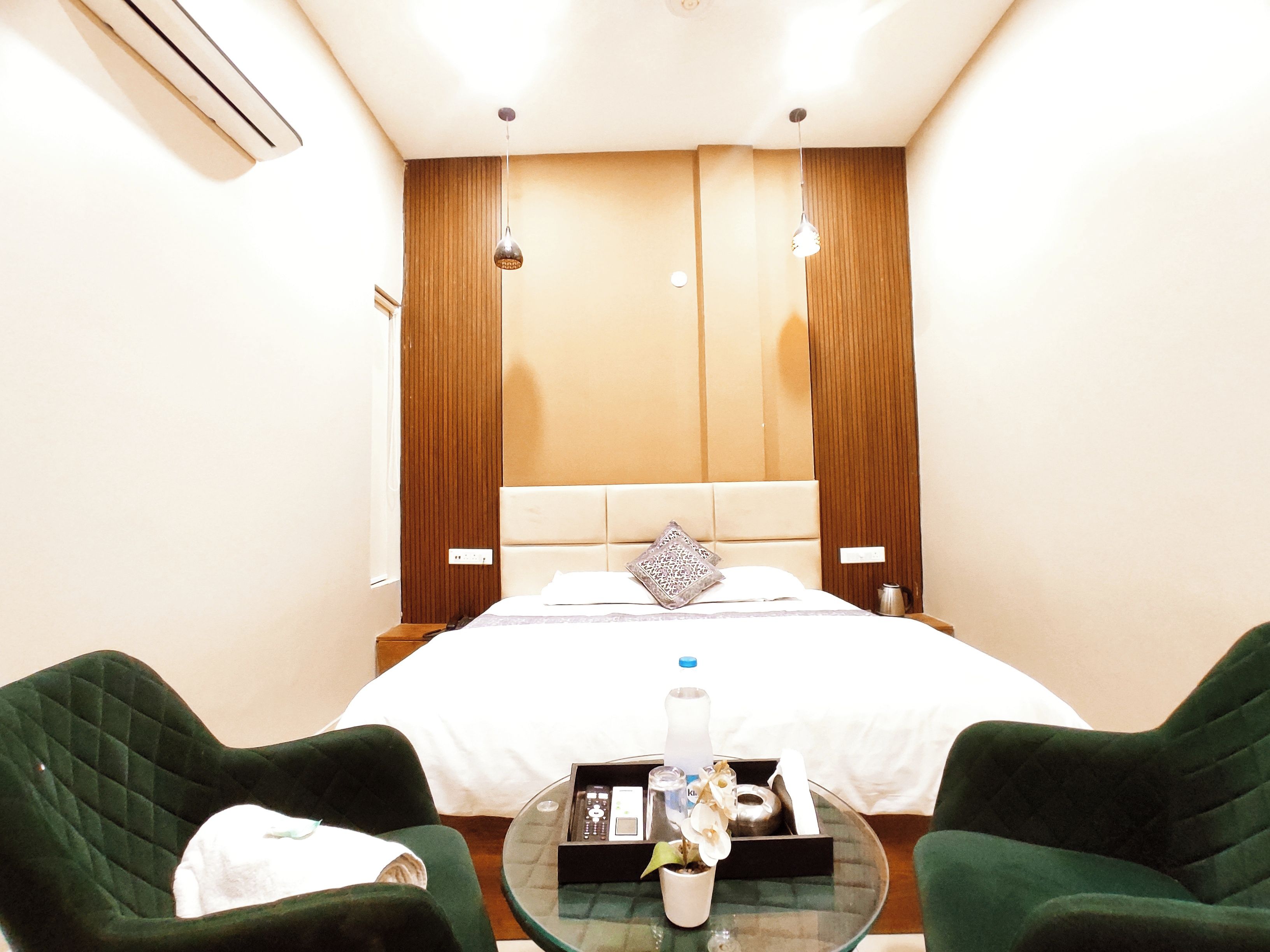 A hotel room with a bed, two green armchairs, a glass coffee table with a bottle and a plant, and a wooden headboard.