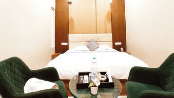A hotel room with a bed, two green armchairs, a glass coffee table with a bottle and a plant, and a wooden headboard.