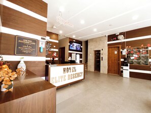 Zona de recepción de un hotel, con mostrador de recepción, televisor y un cartel con el nombre del hotel.