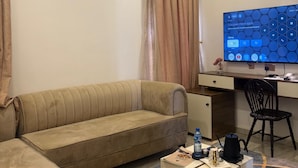 A hotel room with a sofa, a desk with a computer, an air conditioner, and a coffee table with a bottle and a cup. - Merat Hotel And Suites (Jimeta)