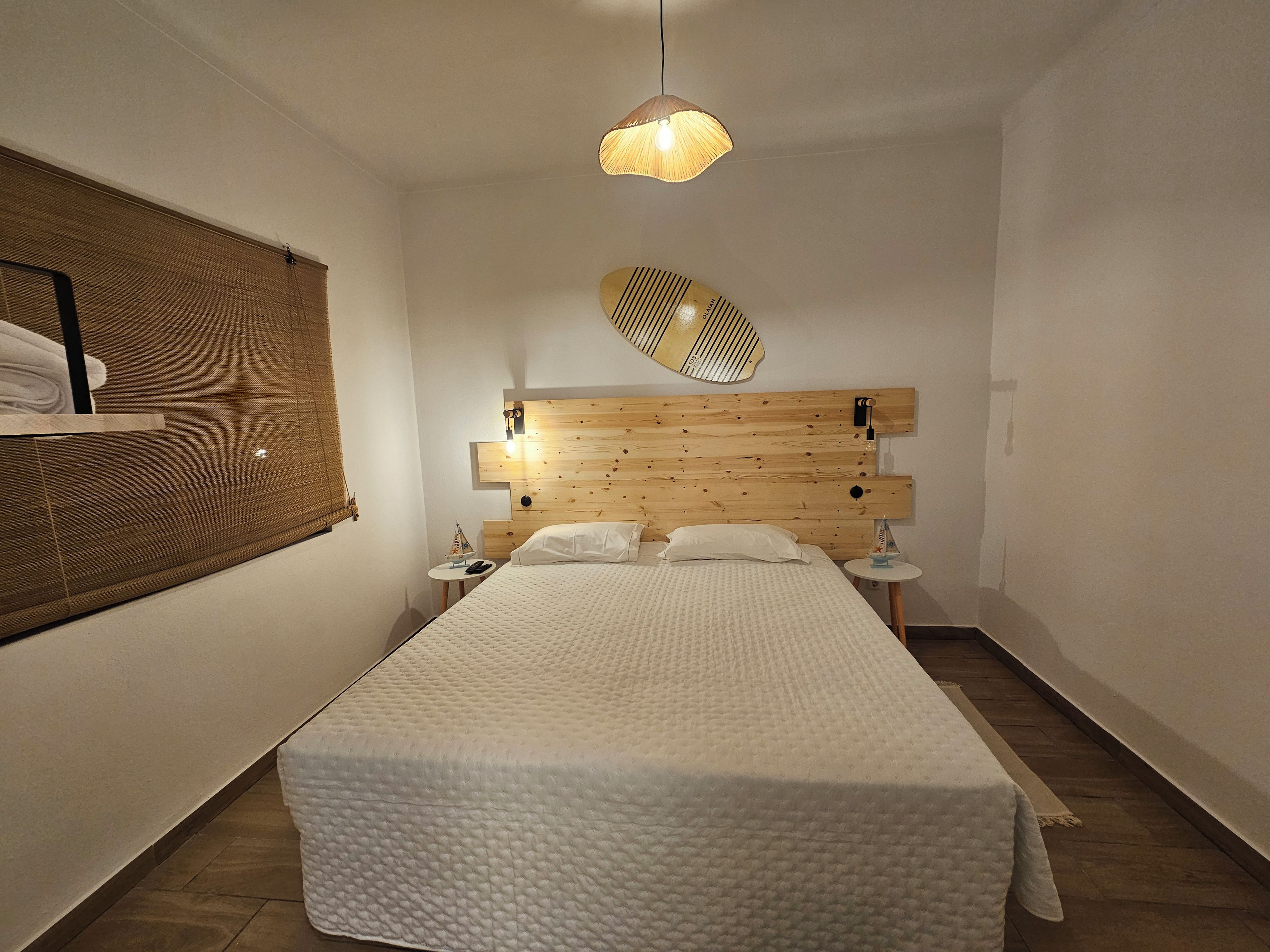Un dormitori amb un panell de fusta al capçal, un llit amb roba de llit blanca, una petita taula de nit amb una làmpada i una estantanya penjada a la paret.