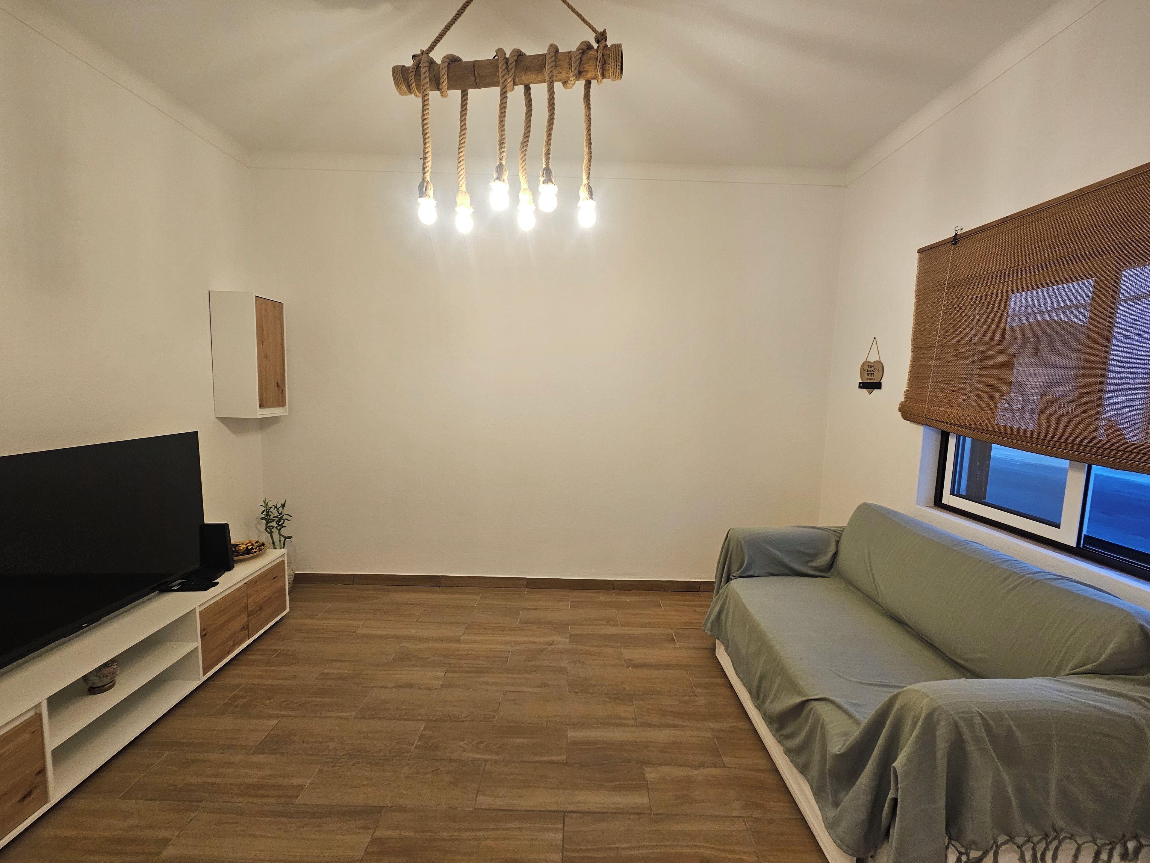 A living room with a flat-screen TV, a wooden floor, a sofa, a hanging light fixture, and a window with blinds.