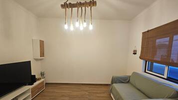 A living room with a flat-screen TV, a wooden floor, a sofa, a hanging light fixture, and a window with blinds.