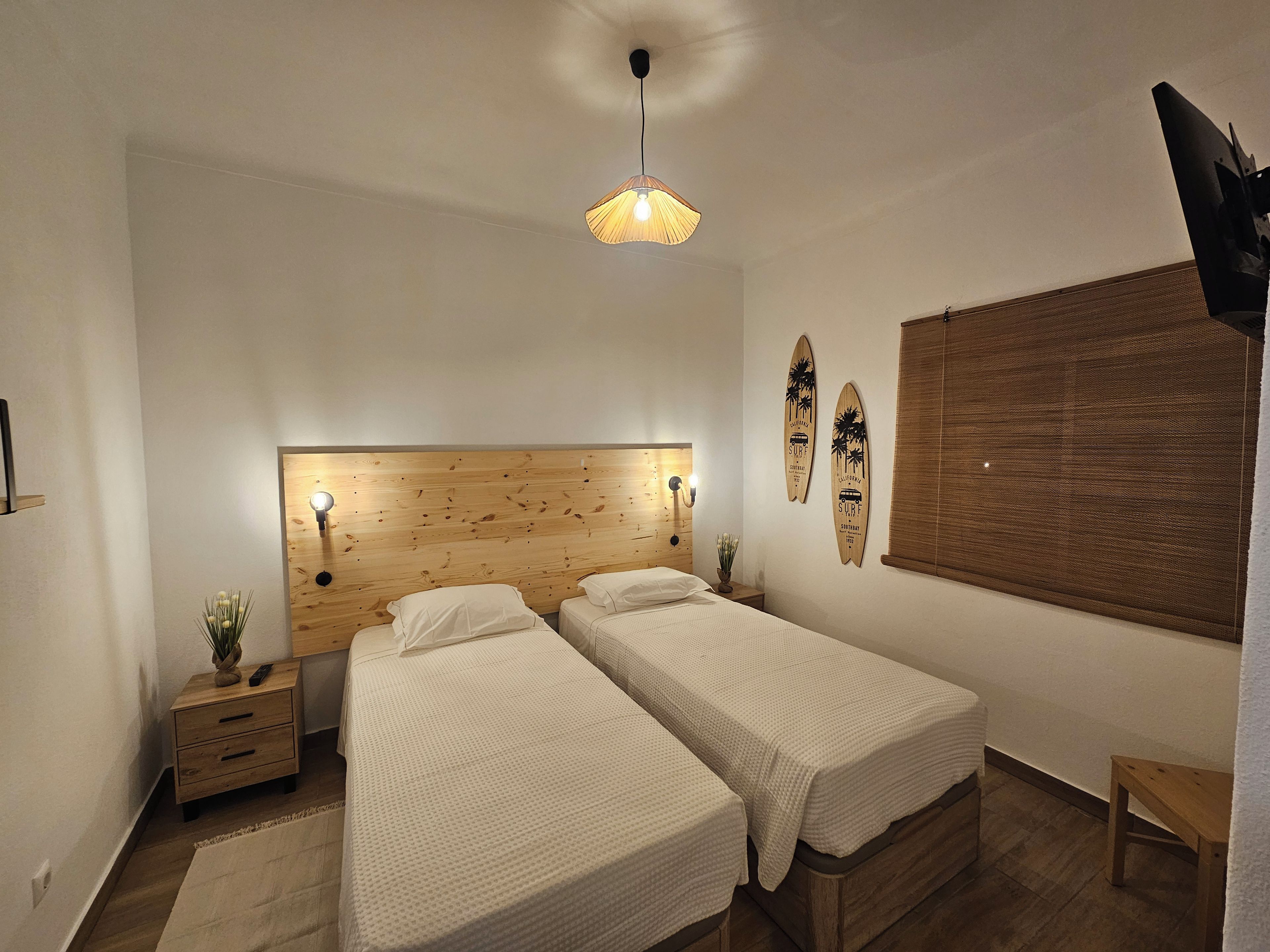 Ein Schlafzimmer mit zwei Betten, einem Holzkopfende, einer Wanddekoration aus einem Surfbrett, einem kleinen Nachttisch mit einer Pflanze und einer Pendelleuchte.