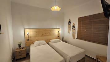 A bedroom with two beds, a wooden headboard, a wall-mounted surfboard decoration, a small bedside table with a plant, and a hanging light fixture.