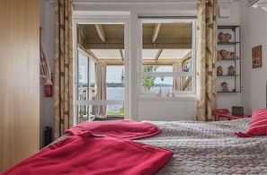 A bedroom with a bed, a window with curtains, a view of a body of water, and a shelf with decorative items. - Lakeside Retreat on Lake Laxsjön – Lake Views, Kayaks & Private Heated Terrace (Dals Långed)
