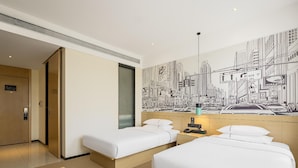 Habitació d'hotel amb dos llits, un mural de paisatge urbà, una porta i una petita taula de nit amb una làmpada.