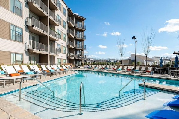 Una piscina con sillones de descanso y un edificio de varios pisos con balcones.