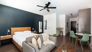Moderni hotellihuone, jossa on sänky, harmaa sohva, ruokailutila pöytineen ja tuoleineen sekä kattotuuletin.