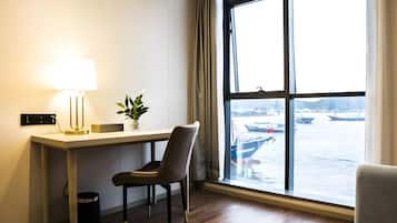 Quarto de hotel com escrivaninha, cadeira, luminária, planta em vaso e uma grande janela com vista para barcos e água.