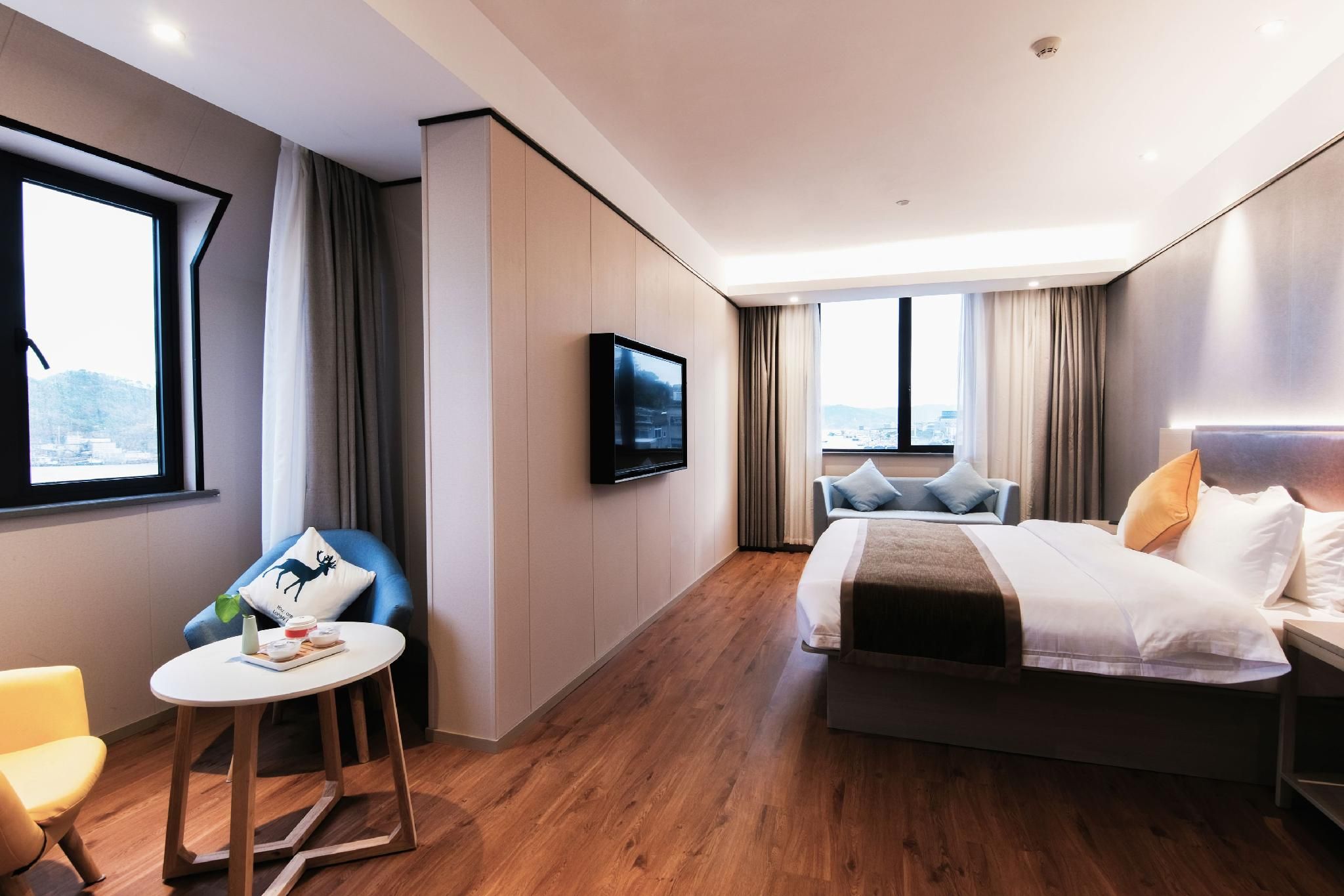 A modern hotel room with a large bed, a seating area with a table, a flat-screen TV mounted on the wall, and a city view through the windows.