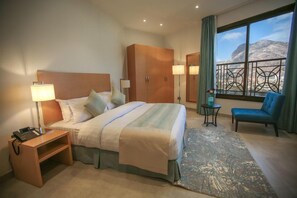 A hotel room with a bed, bedside table, lamp, wardrobe, chair, and a view of mountains through a balcony.
