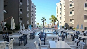 Zona de piscina amb taules i caires, un pont i edificis alts al fons.