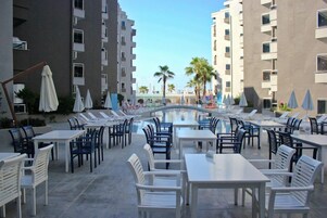 Zona junto a la piscina con mesas y sillas, un puente y edificios altos al fondo.