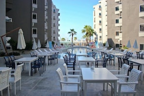 Zona de piscina amb taules i caires, un pont i edificis alts al fons.
