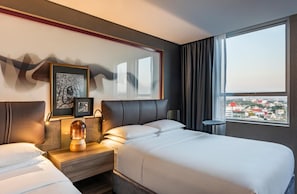 Hotelkamer met een bed, nachtkastjes, een lamp, ingelijste foto's en uitzicht op een stadsbeeld door een raam.