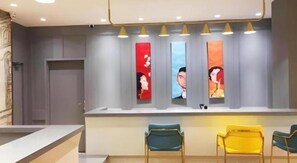 Zona de recepción con mostrador, tres sillas y una fila de obras de arte verticales en la pared.