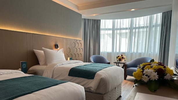 Habitación de hotel con dos camas, un área de descanso con sillas, una mesa y un ventanal con cortinas.