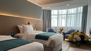 Habitación de hotel con dos camas, un área de descanso con sillas, una mesa y un ventanal con cortinas.