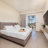 Habitación de hotel con cama, televisión, balcón con mesa y sillas, y vista a palmeras.