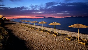 Una playa con sombrillas y sillas de playa, un litoral rocoso y una puesta de sol sobre el agua.