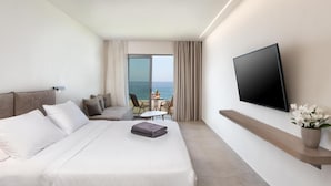 A modern hotel room with a large bed, a flat-screen TV mounted on the wall, a small table with chairs, and a view of the sea through the window. - Ixia Dream - Adults Only (Rhodes)