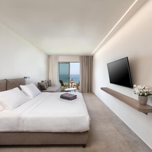 Quarto de hotel moderno com uma cama grande, televisão de tela plana na parede, uma mesa pequena com cadeiras e vista para o mar pela janela.