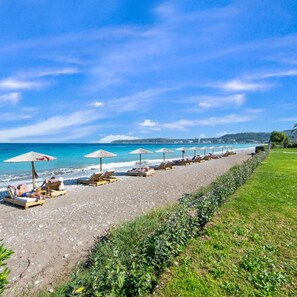 A beach with rows of sun loungers and umbrellas, a grassy area beside the beach, and a clear view of the ocean. - Ixia Dream - Adults Only (Rhodes)