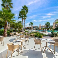 Un área de comedor al aire libre con mesas y sillas, una piscina y palmeras.