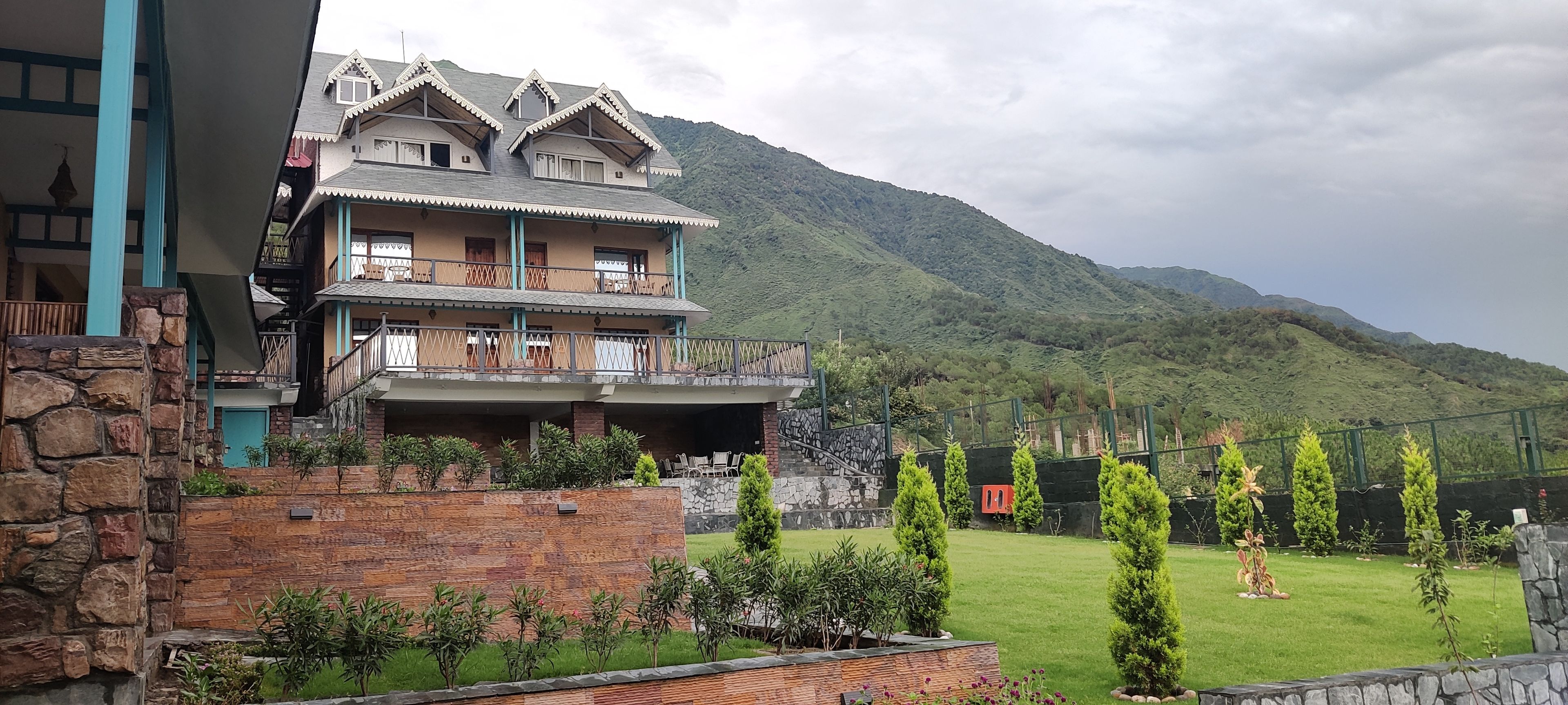 Um edifício com várias andares e varandas, rodeado por um jardim bem cuidado e uma paisagem montanhosa.