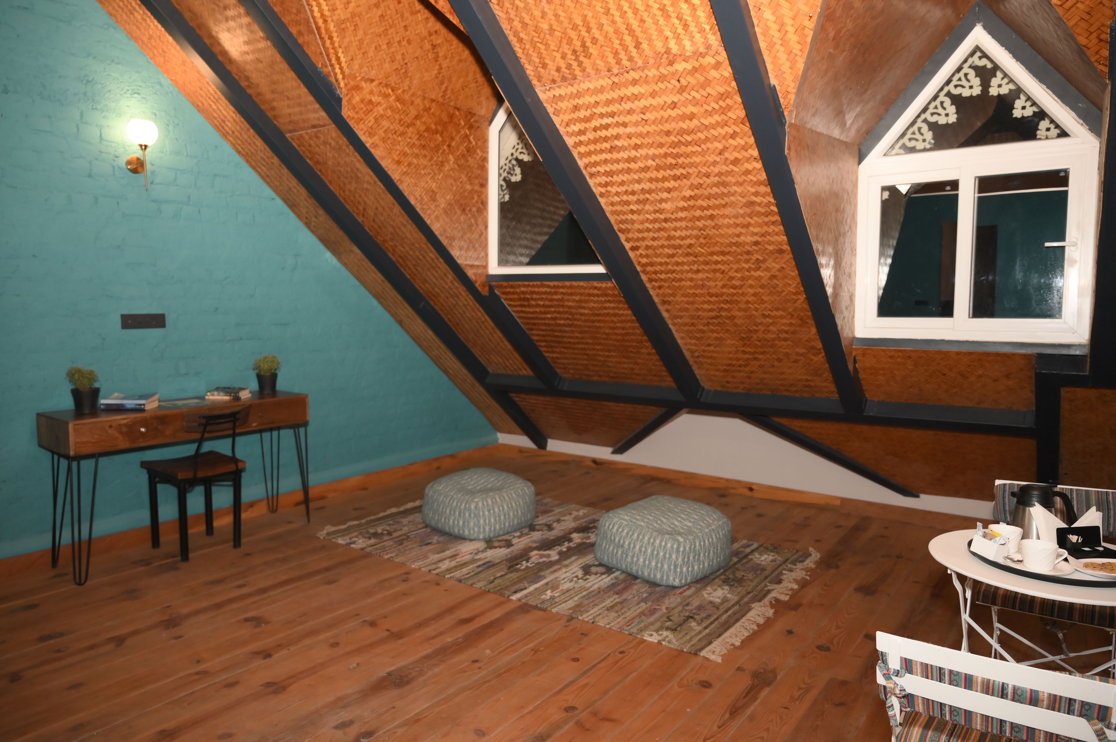 Una habitación con techo inclinado, un escritorio de madera con silla, dos cojines, una ventana y una mesa con hervidor y tazas.