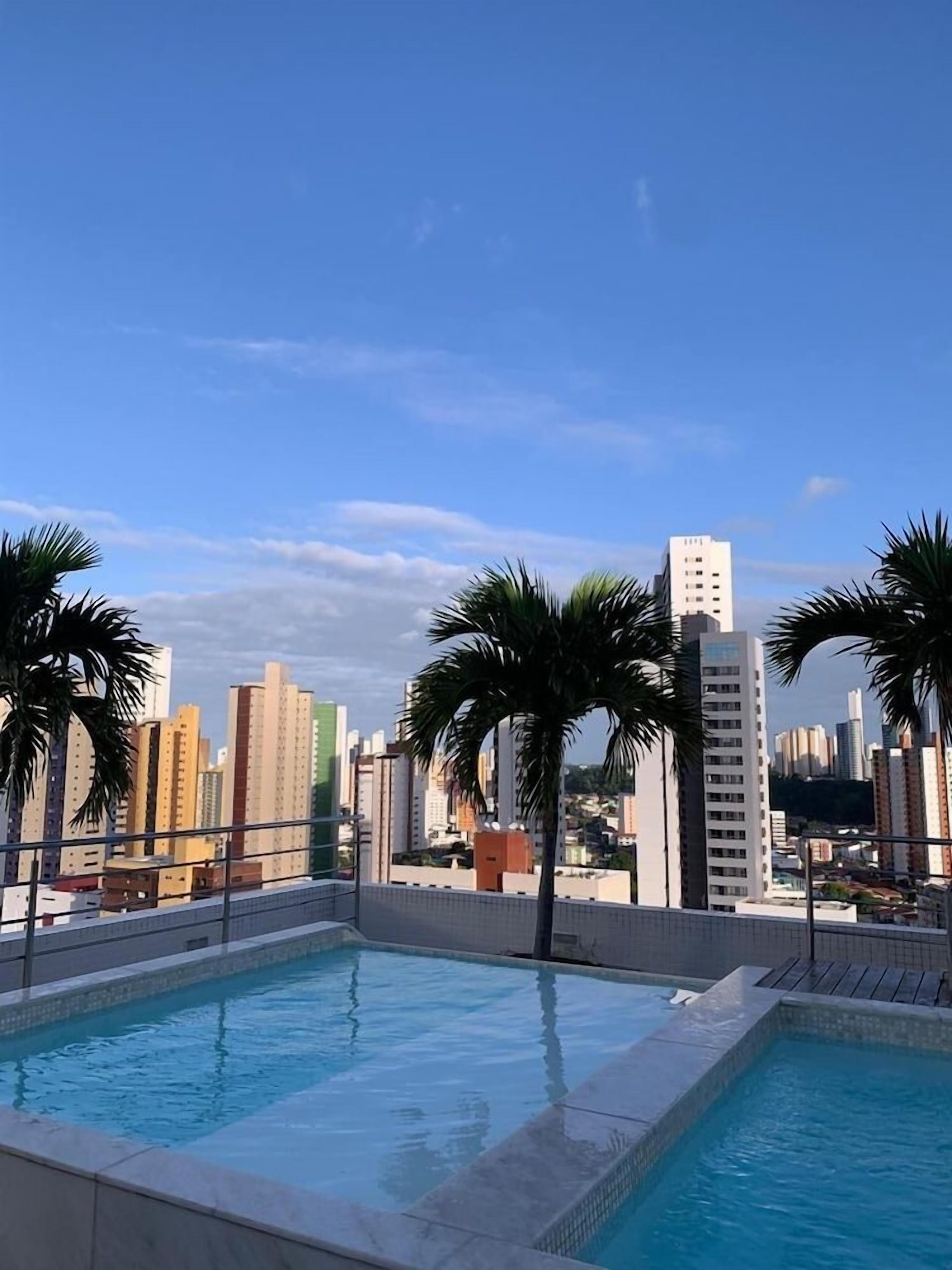 Piscina no terraço com vista para a cidade, cercada por palmeiras.