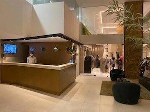 Zona de recepción de un hotel con mostrador, pantalla de televisión y una gran planta en maceta.