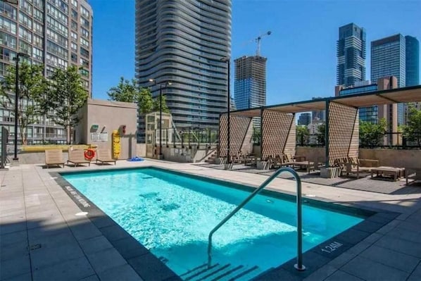 Una piscina con una escalera metálica, rodeada de sillas de descanso y una zona de estar cubierta, con rascacielos al fondo.